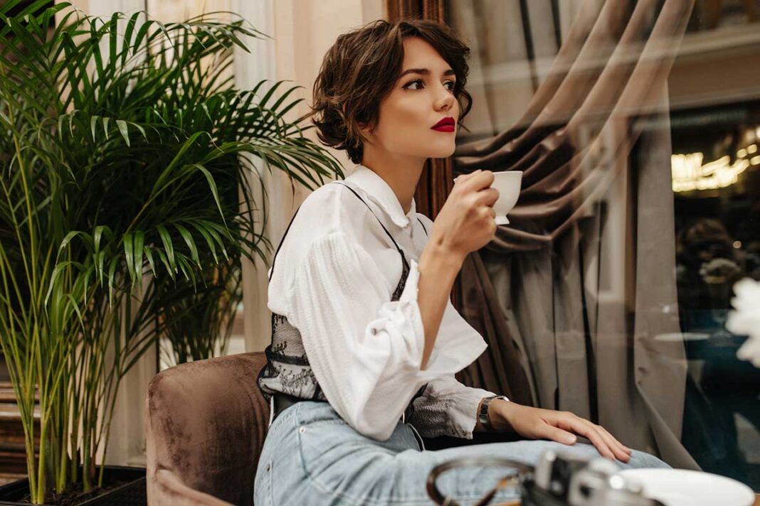 young-woman-with-bright-lips-curly-hair-poses-restaurant-trendy-woman-white-shirt-jeans-holds-cup-coffee-cafe žena pije kavu