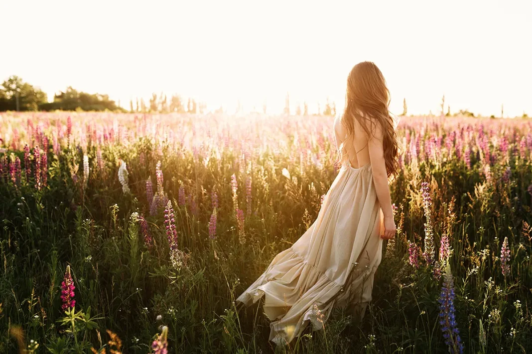 young-woman-walking-flower-field-sunset-background mlada zenska osoba u dugoj haljini na livadi punoj cvijeća